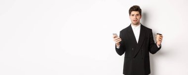 Kafası karışmış bir iş adamının elinde bir fincan kahve ve cep telefonuyla beyaz arka plana karşı duran bir şeyi anlamadığı görüntüsü..