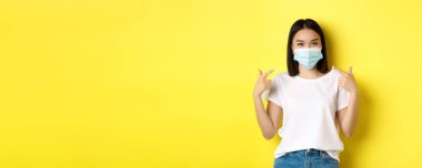 Covid-19, karantina ve sosyal uzaklık kavramı. Beyaz tişörtlü genç Asyalı kadın, Coronavirus 'taki tıbbi maskesini işaret ediyor, sarı arka plan..