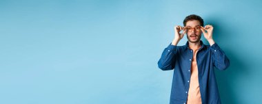 Yakışıklı beyaz bir adamın gözlük takıp kameraya baktığı, gözlükçüde yeni gözlük denediği, mavi arka plan..