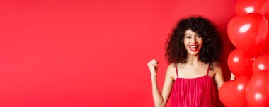 Heyecanlı güzel bir kadın kazanıyor, evet diyor ve gülümsüyor, başarıyı kutluyor, yumruk pompasını motive ediyor, kalp balonları ve kırmızı arka planda duruyor..