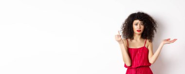 Kıvırcık saçlı üzgün kız arkadaş, yüzük takmayan parmağını gösteriyor ve kafası karışmış görünüyor, kararsız erkek arkadaşından şikayetçi, evlenmek istiyor, beyaz arka planda duruyor..