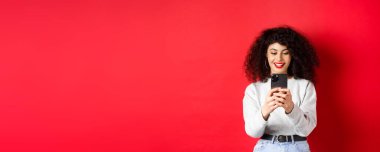Gülümseyen modern kız akıllı telefondan fotoğraf çekiyor, ekrana bakıyor ve video kaydediyor, kırmızı arka planda duruyor..