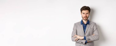 Profesyonel gibi görünen kendine güvenen genç adam, göğsünde çapraz kollar, beyaz arka planda duruyor..