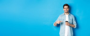 Gülümseyen mutlu adam kahve ile kağıt bardağı işaret ediyor, kafe tavsiye ediyor, mavi arka planda duruyor..