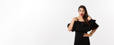 Moda ve güzellik. Şok olmuş bir kadın kendini gösteriyor ve kameraya bakıyor, suçlanıyor, beyaz arka planda siyah elbiseyle dikiliyor..