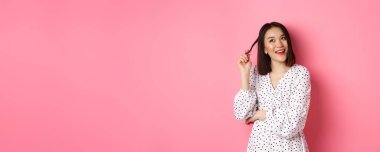 Hayalperest Asyalı kız düşünüyor, alışveriş yapıyor ve gülümsüyor, sol üst köşeye bakıyor düşünceli bir gülümsemeyle, pembe arka plan üzerinde duruyor..