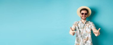 Yaz şapkalı ve Hawaii gömlekli mutlu bir turist. Ortadaki logoyu işaret ediyor. Bir şey gösteriyor, mavi arka planda duruyor..