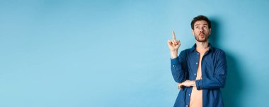 Yakışıklı bir adamın bir fikir sunduğu, parmağını kaldırdığı ve mavi arka planda durup düşünerek kafasını kaldırdığı bir görüntü. Boşluğu kopyala
