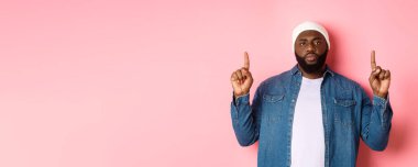 Ciddi ve canı sıkkın sakallı siyahi adam, kameraya bakıyor ve işaret ediyor, reklamını yapıyor, pembe arka planda duruyor..