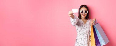 Güneş gözlüklü güzel Asyalı kadın alışverişe çıkıyor, çantalarını tutuyor ve kredi kartını gösteriyor, pembe arka planda duruyor..