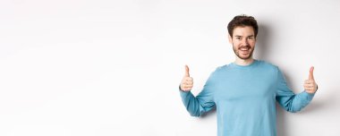 Neşeli genç adam başparmağını kaldırıyor ve gülümsüyor, iyi ürün öneriyor, seçimi övüyor, beyaz arka planda duruyor..