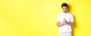 Akıllı telefon ekranından rahatsız olmuş gibi görünen, telefonda garip mesajlar okuyan, beyaz tişörtlü, sarı arka planda duran bir adam..
