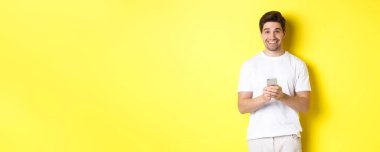 Akıllı telefondaki tanıtım teklifini okuduktan sonra gülümseyen ve mutlu görünen bir adam beyaz tişörtlü sarı arka planda duruyor..