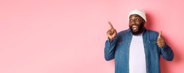 Mutlu Afro-Amerikalı adam baş parmağını yukarı kaldırıyor ve sol üst köşedeki fotokopi alanını işaret ediyor, iyi bir anlaşmaya övgüler yağdırıyor, pembe arka planda duruyor..