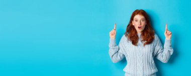 Kış tatili ve insanların konsepti. Şaşkın kızıl saçlı genç kız işaret ediyor, reklam veriyor ve kameraya bakıyor, mavi arka plan..