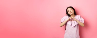 Sevgililer Günü konsepti. Güzel Koreli genç kalp jesti yapıyor ve hayranlıkla gülümsüyor, aşık oluyor, romantik hissediyor, pembe arka plan.