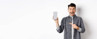 Heyecanlı yakışıklı adam boş akıllı telefon ekranı gösteriyor ve gülümsüyor, şuraya bakın, jest, beyaz arka planda duruyor..