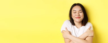 Romantik Asyalı kızın kendine sarılıp hayal kurması, gözlerini kapatması ve gülümsemesi, sarı arka planda durduğunu hayal ederken..