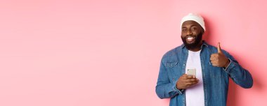 Teknoloji ve online alışveriş konsepti. Mutlu Afro-Amerikalı adam cep telefonu kullanırken onay veriyor, pembe arka planda tatmin olmuş duruyor..