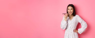 Güzel Koreli kadın hoparlörden konuşuyor, sesli mesaj kaydediyor ve mutlu bir şekilde gülümsüyor, pembe arka planda duruyor..