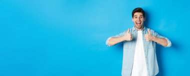 Gülümseyen yetişkin bir adam heyecanlı bir yüz ifadesiyle başparmağını kaldırıyor, harika bir şey gibi, ürünü onaylıyor, mavi arka planda duruyor..