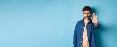 Üzgün adam kaşlarını çatıyor ve durmak istiyor, yasaklamak için elini uzatıyor ya da kötü bir şeye karşı çıkıyor, mavi arka planda duruyor..