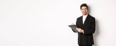 Siyah takım elbiseli yakışıklı, başarılı patronun resmi. Belgeleri imzalarken göz kırpıyor ve gülümsüyor. Beyaz arka planda duruyor..