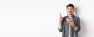 Doğum günü. Mutlu genç adam, pasta ve çapraz parmaklarla dilek diliyor, umut dolu görünüyor, beyaz arka planda duruyor..