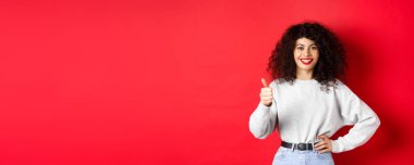 Kıvırcık saçlı, kırmızı dudaklı, tutkulu genç bir kadın. Başparmaklarını gösterip onaylıyor ve gülümsüyor. Kırmızı arka planda duran iyi ürünü övüyor..