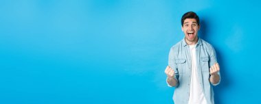 Heyecanlı yakışıklı adam kazanıyor, kendini şanslı ve mutlu hissediyor, zaferi kutluyor ve evet diye bağırıyor, mavi arka planda duruyor..