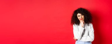 Kırmızı dudaklı, sıkkın ve yorgun kıvırcık kadın, kameraya bakıyor, stüdyo arka planında duruyor..