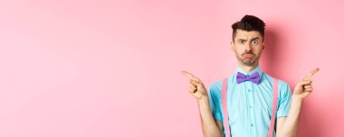 Kararsız genç adam dudaklarını büzüyor, kaşlarını çatıyor, parmaklarını iki şeye doğrultuyor, karar veremiyor, pembe arka plan üzerinde şaşkın şaşkın duruyor..