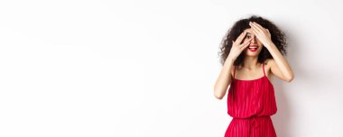 - Sürpriz. Heyecanlı, kıvırcık saçlı, kırmızı dudaklı, parmaklarının arasından bakan, şaşkın bir şekilde gülümseyen, tanıtıma bakan, beyaz arka planda duran güzel kız..