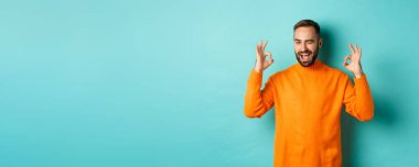 Kendine güveni tam bir adam, tamam işaretleri gösteriyor ve sana göz kırpıyor, ürün öneriyor, stüdyo arka planında turuncu süveter giyiyor..