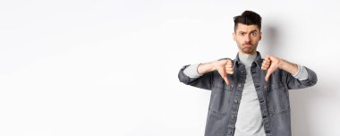 Hayal kırıklığına uğramış komik adam somurtuyor ve üzgün görünüyor, baş parmaklarını gösteriyor hoşnutsuz, beyaz arka planda duruyor..