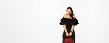 Güzellik ve moda konsepti. Uzun boylu, somurtan, şaşırmış görünen, el çantası tutan, topuklu ve siyah elbise giyen, beyaz arka planlı bir kadın..