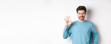 - Tamam. Yakışıklı gülümseyen adam iyi bir işaret veriyor ve kendinden emin görünüyor, iyi bir şey tavsiye ediyor veya övüyor, beyaz arka planda duruyor..