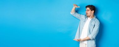 Heyecanlı yakışıklı adam büyük bir nesne gösteriyor ve şaşırmış görünüyor, mavi arka planda duruyor..