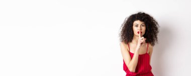 Kıvırcık saçlı güzel bir kadın, kırmızı elbise giyiyor, kameraya susuyor, sırrını söylüyor, sana sus diyor, beyaz arka planda duruyor..