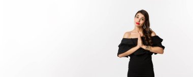 Moda ve güzellik. Güzel, şık bir kadın yardım istiyor, bir şey için yalvarıyor, ellerini birbirine bastırıp, siyah elbise içinde, beyaz arka planda durarak lütfen diyor..