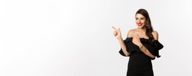 Moda ve güzellik. Mücevher, makyaj ve siyah elbise giymiş çekici bir kadın. Gülen ve parmakla işaret eden beyaz bir arka plan..