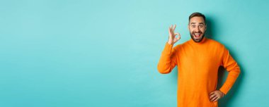 Etkilenmiş bir adam tanıtım öneriyor, tamam işareti gösteriyor ve gülümsüyor. Şaşırmış, onaylamış ve katılıyor, açık mavi arka planda duruyor..