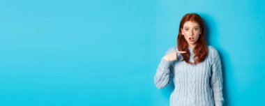 Kafası karışmış kızıl kız kendini işaret ediyor, seçiliyor, mavi arka planda süveter giyiyor..