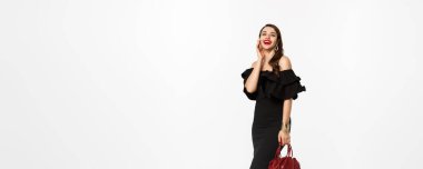 Güzellik ve moda konsepti. Topuklu ayakkabı, siyah elbise ve makyajla partiye giden, zarif bir çanta taşıyan, kameraya gülen, beyaz arka planda güzel bir bayan..