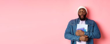Eğitim. Heyecanlı Afro-Amerikalı yetişkin öğrenci elinde not defterleri, kameraya hayranlıkla bakıyor, pembe arka planda duruyor..