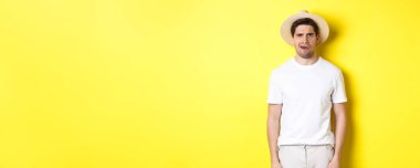 Turizm ve yaz kavramı. Kafası karışmış hasır şapkalı gezgin, şaşkın görünüyor, bir şeyi anlamıyor, sarı arka planda duruyor..