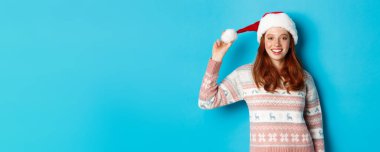 Kış ve Noel arifesi konsepti. Noel Baba şapkalı tatlı kızıl kız kameraya gülümsüyor, mavi sırt grubuna karşı süveter giyiyor..