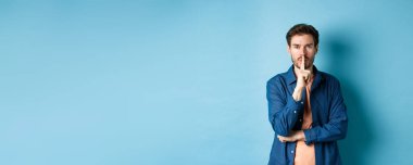 Modern adam kameraya şşş yapıyor, parmaklarını dudaklarına bastırıyor ve bir sır söylüyor, tabu işareti yapıyor, mavi arka planda duruyor..