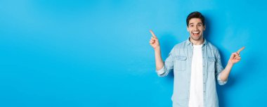 Neşeli yetişkin adam gülümsüyor, parmaklarını yana doğru gösteriyor, sol ve sağ reklam pankartlarını gösteriyor, mavi arka planda duruyor..