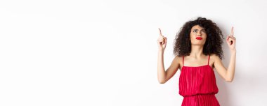 Kızgın ve huysuz bir kadın, kıvırcık saçlı, kırmızı elbiseli, kaşlarını çatmış, hayal kırıklığına uğramış, beyaz arka planda duruyor..